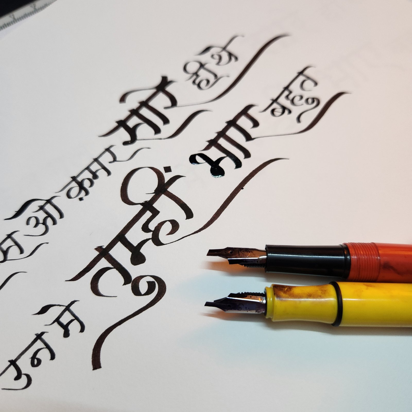 Devanagari letterforms written with broad nib fountain pens, shown beside the pens